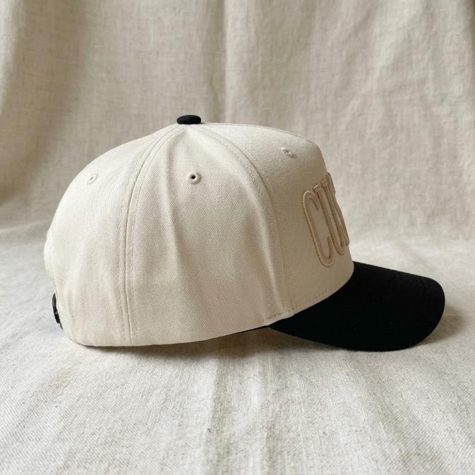 Side view of the ivory trucker hat with black bill and embroidered lettering