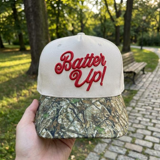 Two-tone baseball trucker hat with red Batter Up embroidery and camouflage bill held outdoors