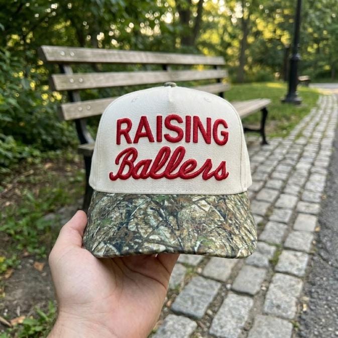 Raising Ballers hat with red embroidered lettering and camouflage-patterned bill held outdoors