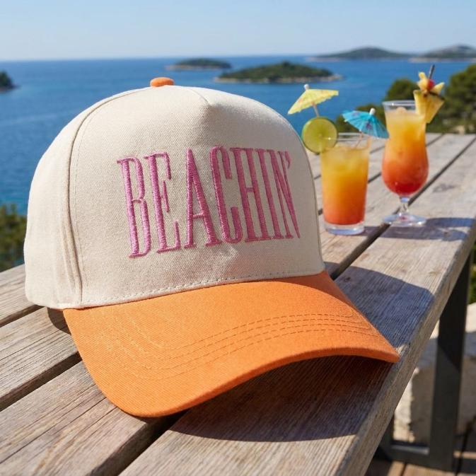 Cream and orange Beachin' hat with pink front embroidery displayed on a wooden table by the sea