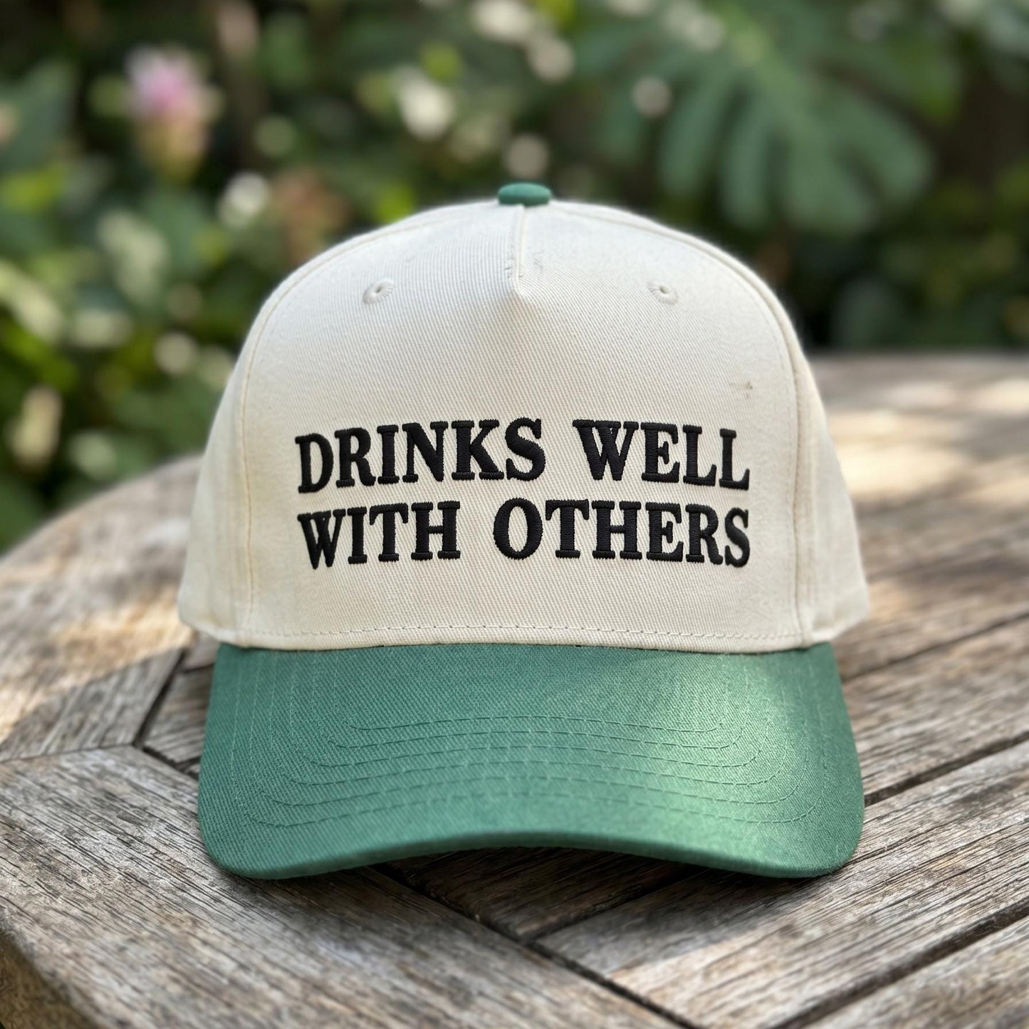 Drinks Well With Others embroidered trucker cap displayed outdoors on a wooden table