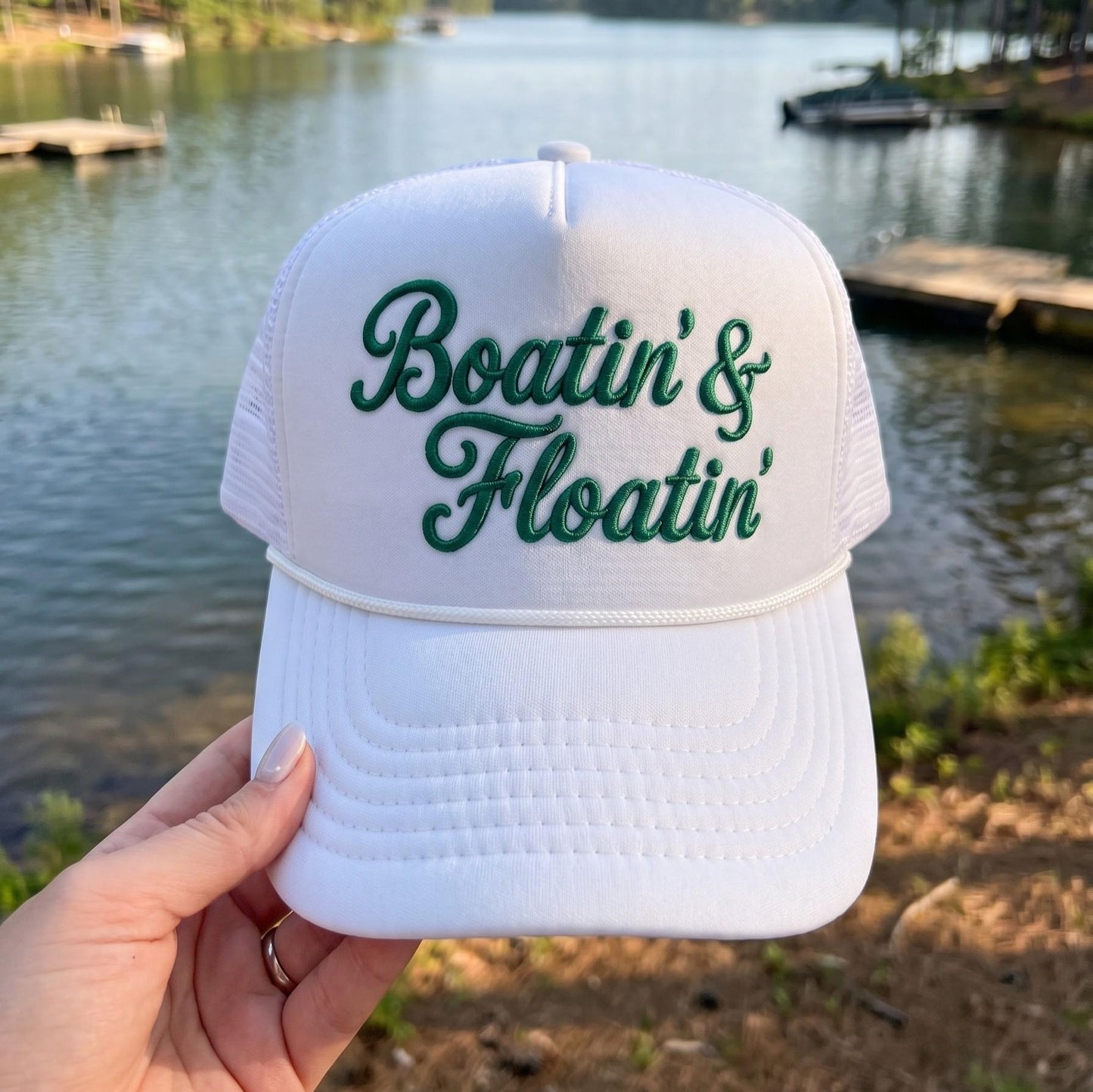 White Boatin' & Floatin' embroidered trucker hat beside a lake