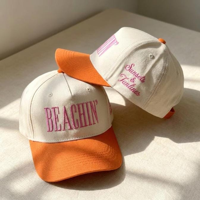 Two cream and orange two-tone hats with pink Beachin' embroidery displayed in warm sunlight