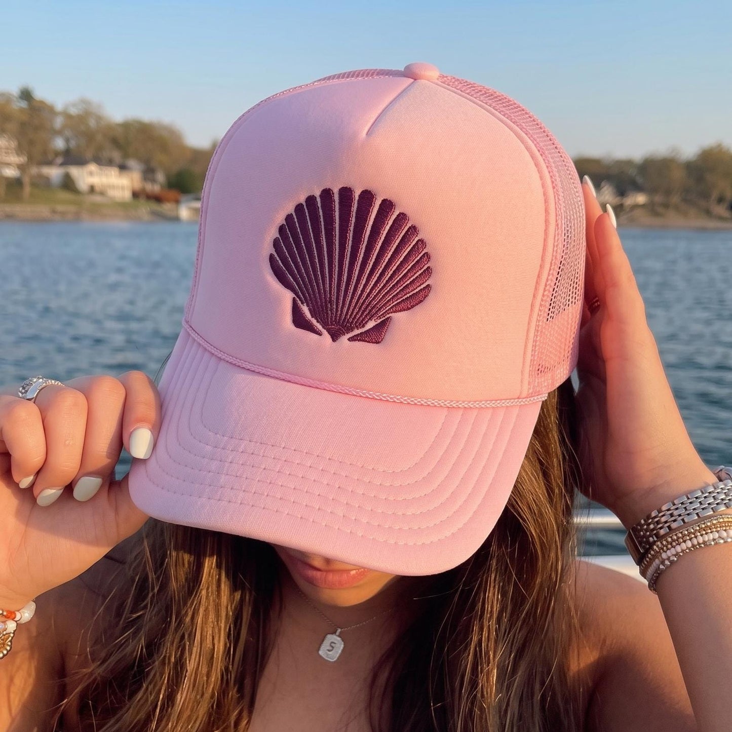 Pink trucker hat with embroidered seashell motif held beside a waterfront