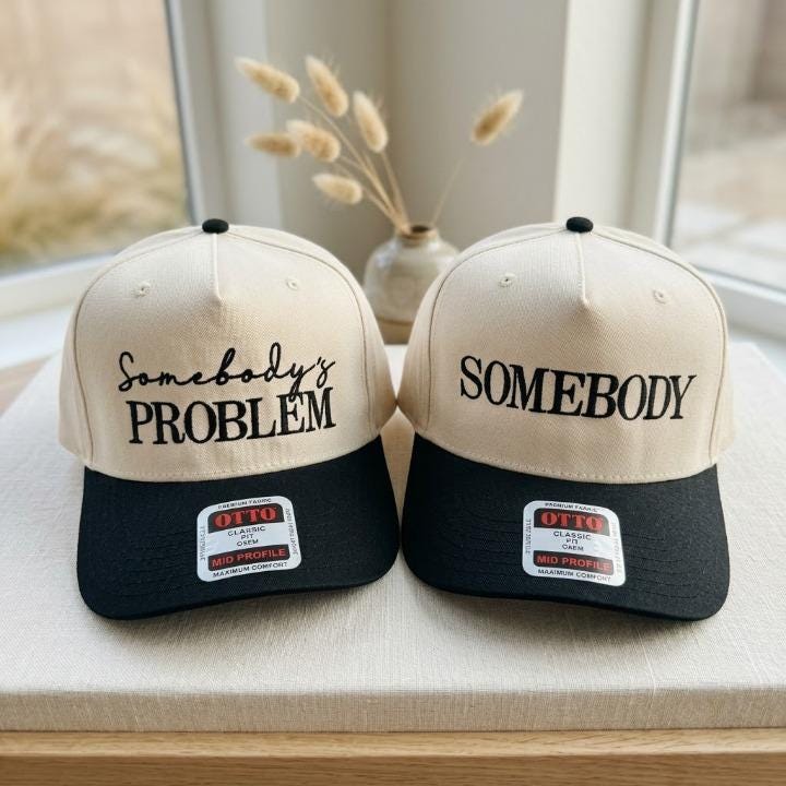 Two two-tone embroidered trucker hats with cream crowns and black bills featuring Somebody’s Problem and Somebody lettering