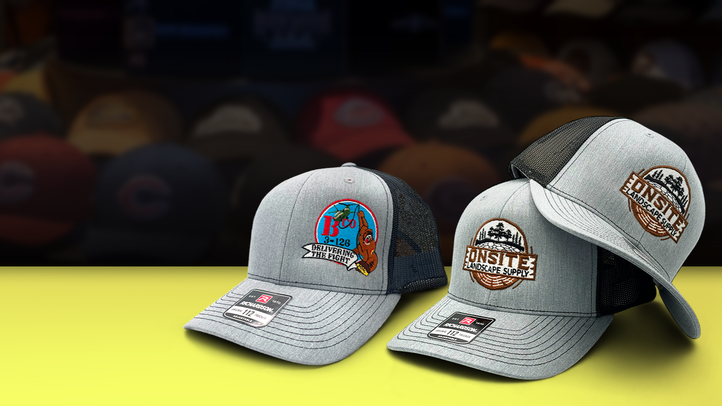 Two gray baseball caps with patches on a yellow surface, blurred background of more caps.