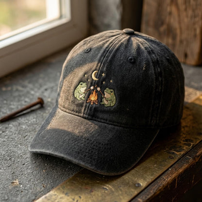 Black embroidered hat featuring two frogs around a campfire beneath a crescent moon and stars