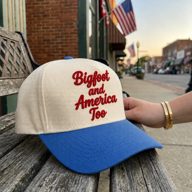 Two-tone Bigfoot and America Too embroidered trucker cap resting on a wooden bench