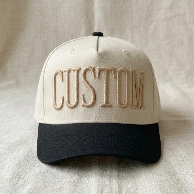 Front view of an ivory and black two-tone trucker hat with raised CUSTOM embroidery