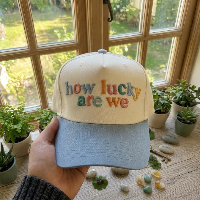 Two-tone embroidered trucker cap with colorful “how lucky are we” lettering and a light blue brim