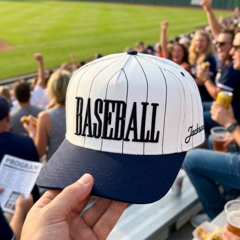 Pinstripe baseball hat with white crown, navy bill, and black BASEBALL lettering held at a baseball game