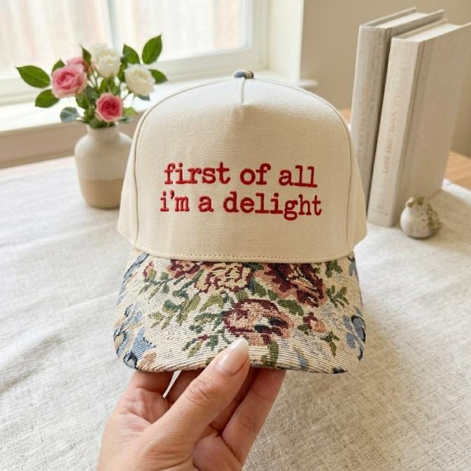 Front view of cream embroidered hat with red “first of all i’m a delight” lettering and floral-patterned brim