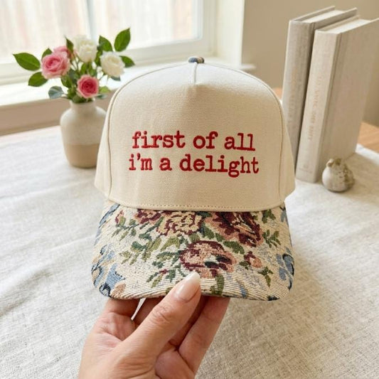 Front view of cream embroidered hat with red “first of all i’m a delight” lettering and floral-patterned brim