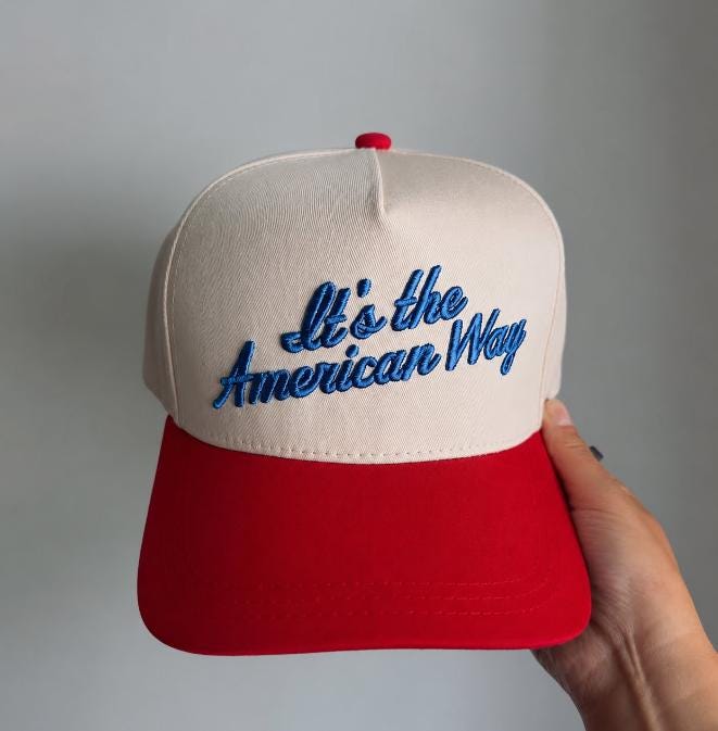 Cream and red two-tone trucker hat with blue embroidered “It’s the American Way” lettering