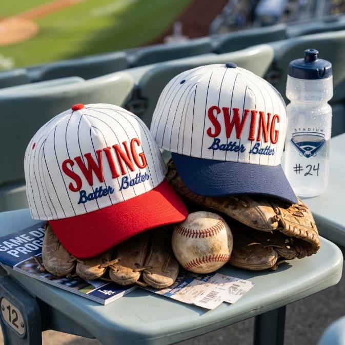 Red and navy pinstripe SWING Batter Batter hats displayed with baseball gloves and a baseball in stadium seating.