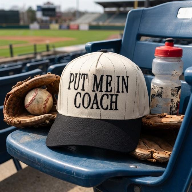 Put Me In Coach baseball hat displayed on a stadium seat beside baseball gloves and a ball