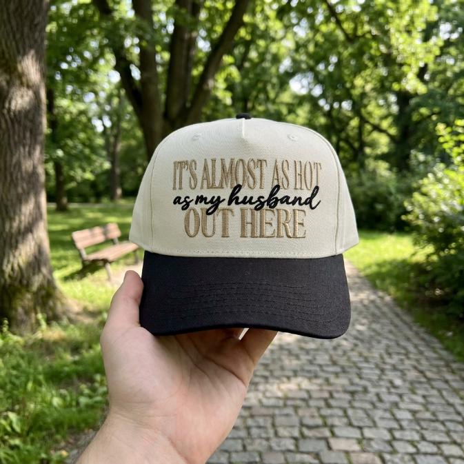 Two-tone trucker hat embroidered with the phrase It’s almost as hot as my husband out here