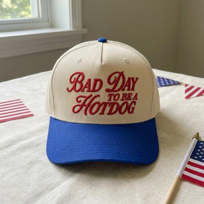 Front view of a cream and blue two-tone trucker cap with red “Bad Day to Be a Hotdog” embroidery