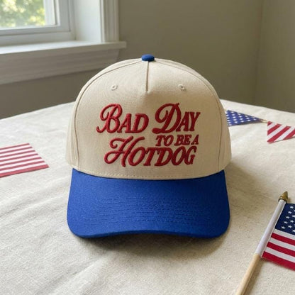 Front view of a cream and blue two-tone trucker cap with red “Bad Day to Be a Hotdog” embroidery