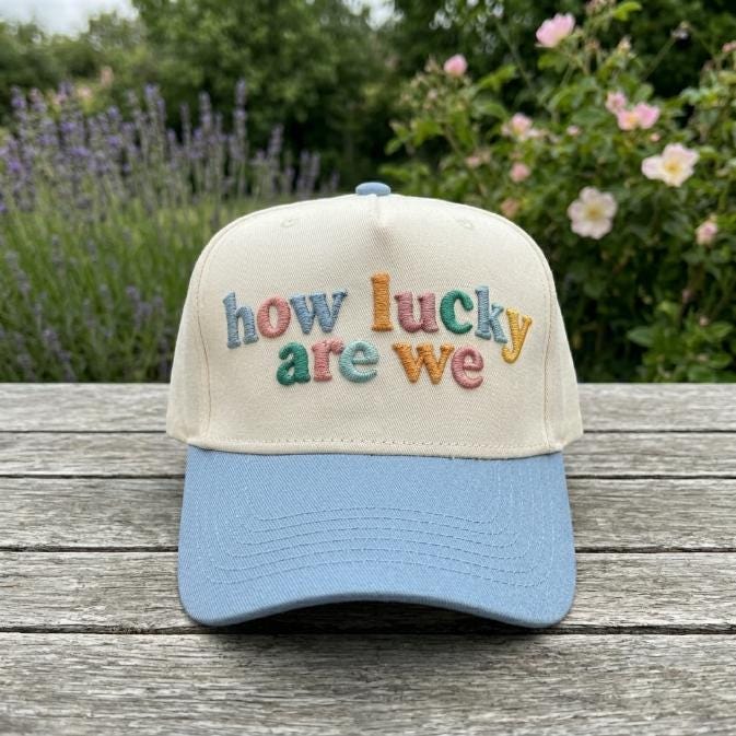 Front view of a cream and light blue “how lucky are we” embroidered trucker cap outdoors