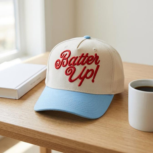 Two-tone trucker cap with red “Bater Up!” embroidery and light blue bill on a wooden table