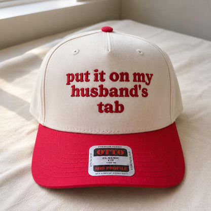Cream and red two-tone trucker hat with red embroidered “put it on my husband’s tab” lettering