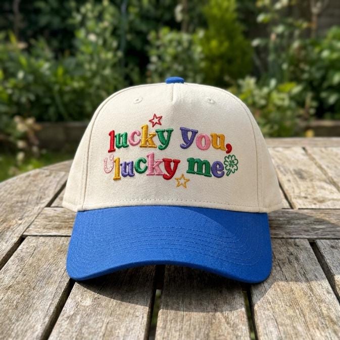 Front view of a cream and blue two-tone hat with colorful Lucky You Lucky Me embroidery, stars, and a four-leaf clover