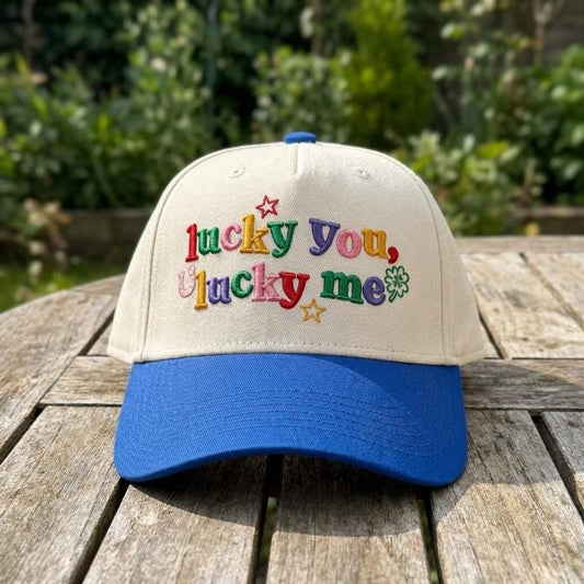 Front view of a cream and blue two-tone hat with colorful Lucky You Lucky Me embroidery, stars, and a four-leaf clover