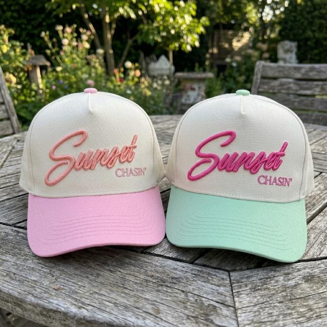 Sunset Chasin' embroidered trucker caps in pink and mint green on a garden table