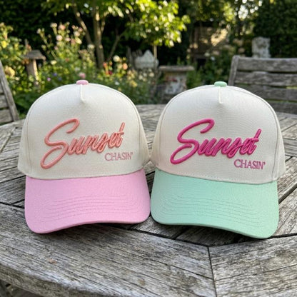 Sunset Chasin' embroidered trucker caps in pink and mint green on a garden table