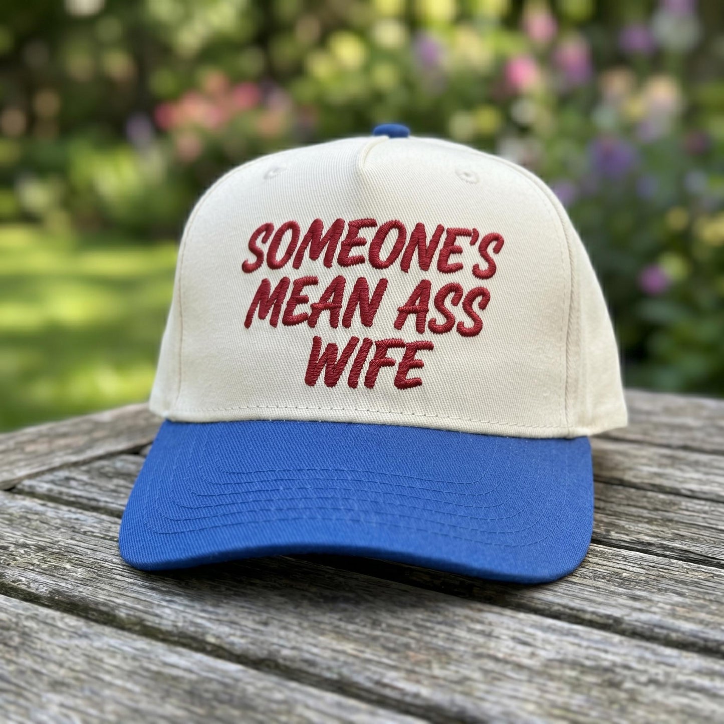 Cream and blue embroidered Someone’s Mean Ass Wife trucker hat displayed outdoors