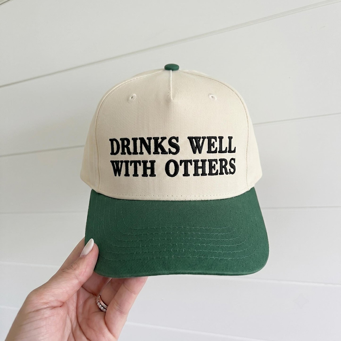 Two-tone cream and forest green trucker hat embroidered with Drinks Well With Others