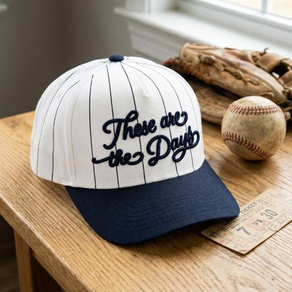 White pinstripe baseball hat with raised navy “These Are the Days” script embroidery on a wooden table