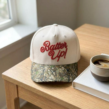 Batter Up embroidered baseball hat with camo bill displayed beside coffee and a book