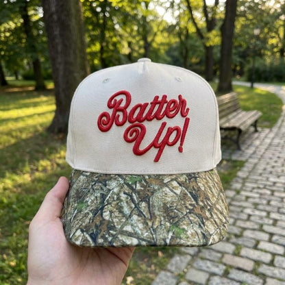 Two-tone baseball trucker hat with red Batter Up embroidery and camouflage bill held outdoors