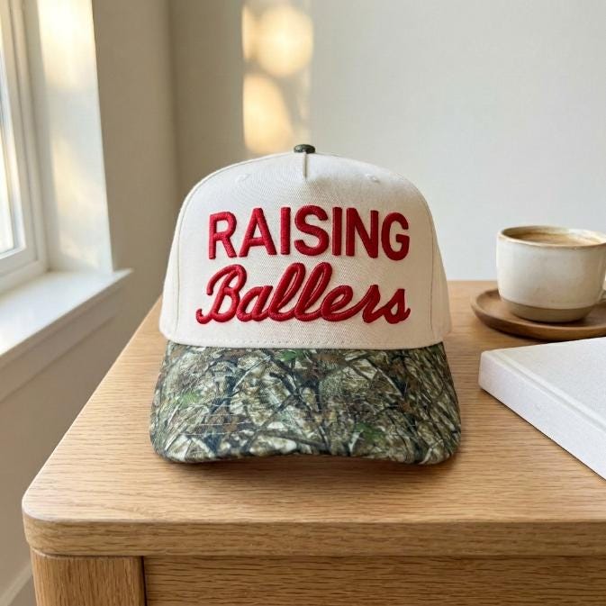 Raising Ballers embroidered hat displayed on a wooden table beside a coffee cup