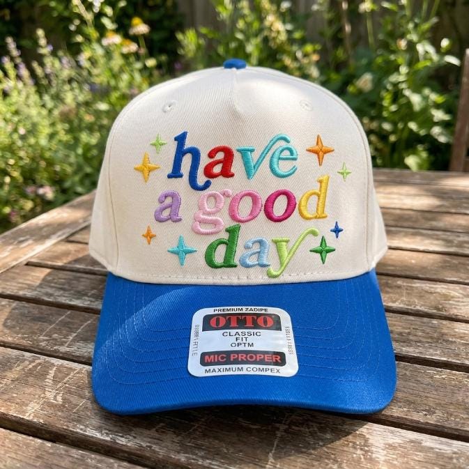 White and blue embroidered trucker hat with colorful “have a good day” lettering and star accents