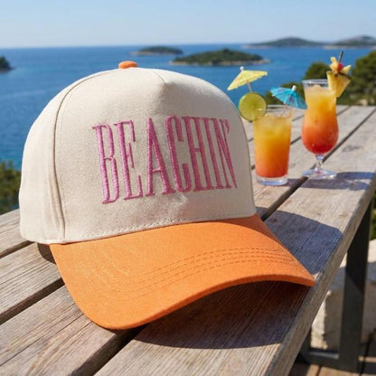 Cream and orange Beachin' hat with pink front embroidery displayed on a wooden table by the sea