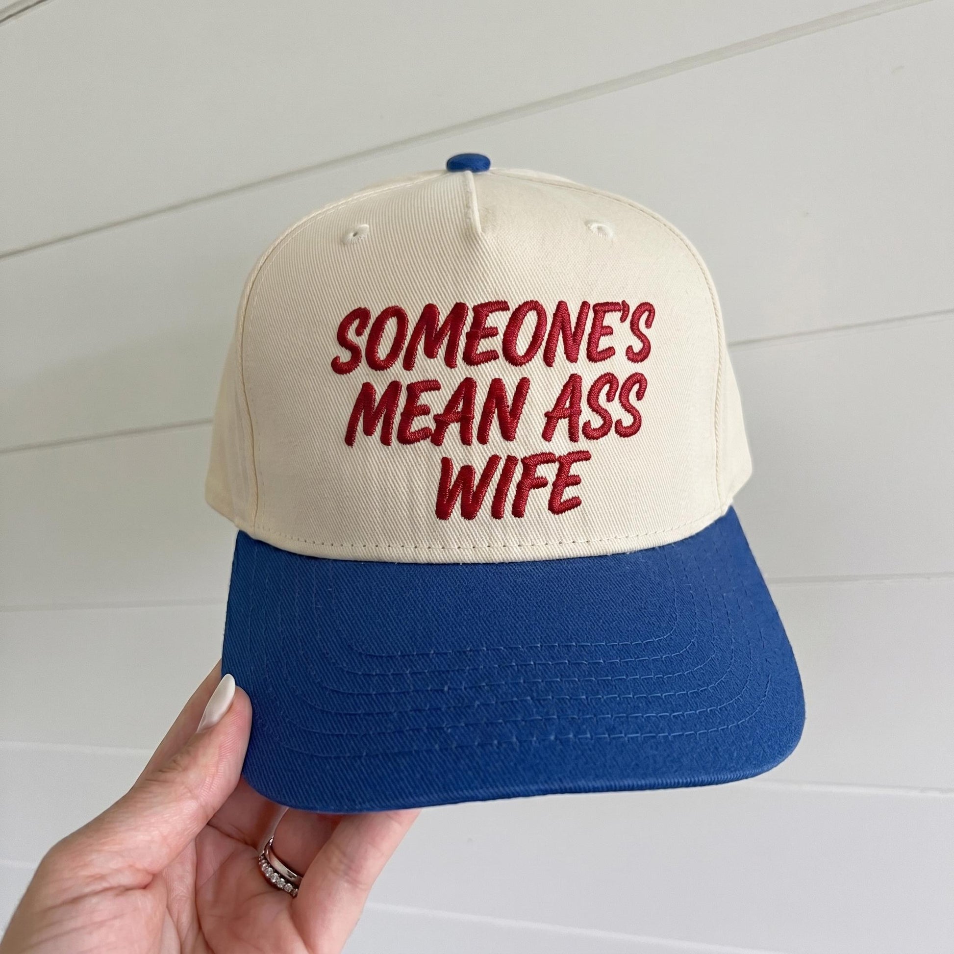 Cream and blue trucker hat with red embroidered Someone’s Mean Ass Wife text held in hand