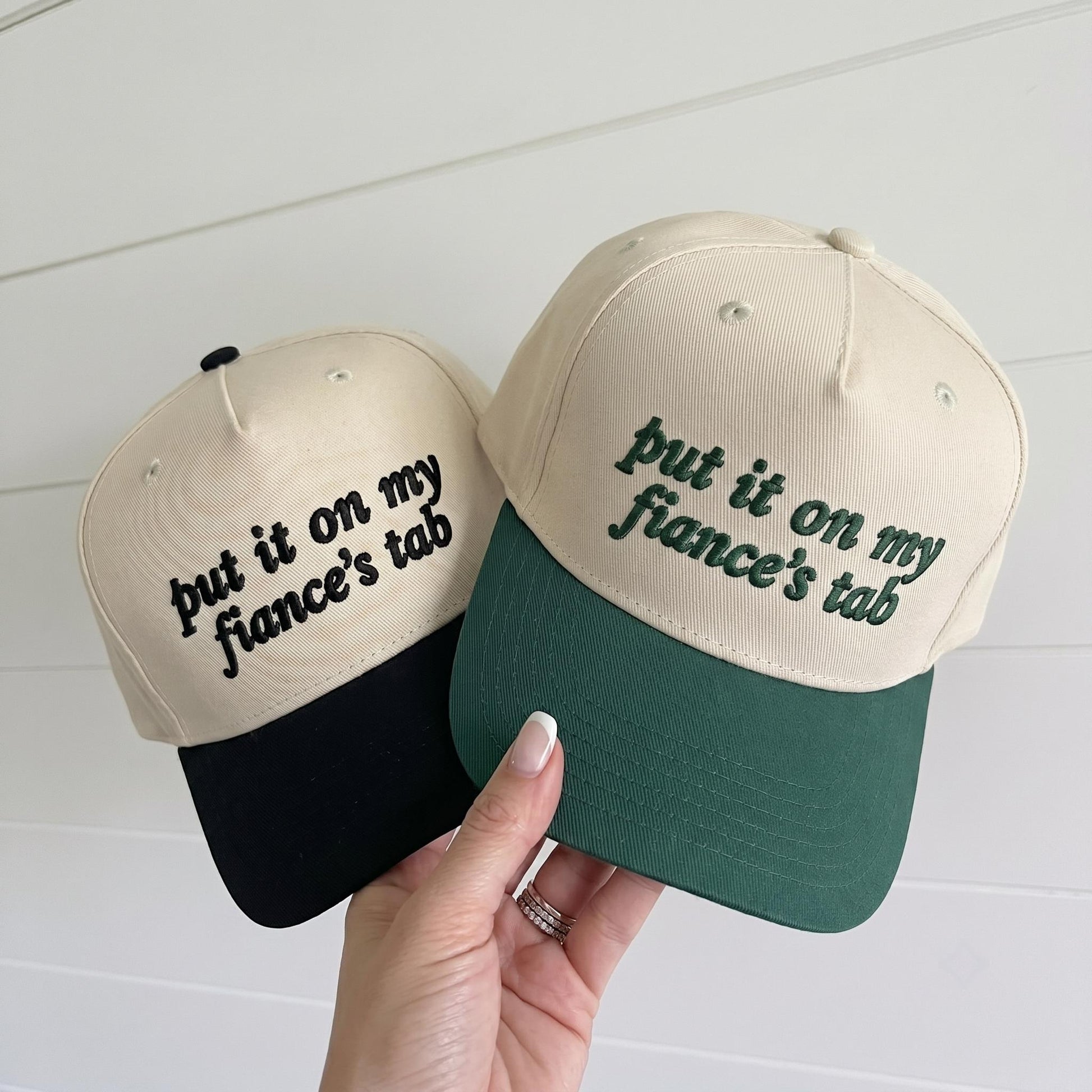 Two embroidered two-tone trucker hats reading put it on my fiance's tab in black and green brim styles