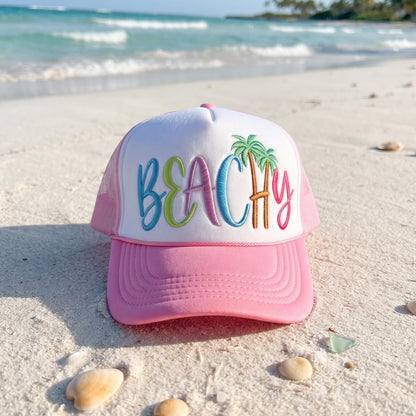 Pink beach trucker hat with embroidered BEACH lettering photographed on a sandy beach