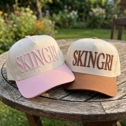 Pink and brown SKINGRL embroidered trucker caps displayed outdoors on a wooden table