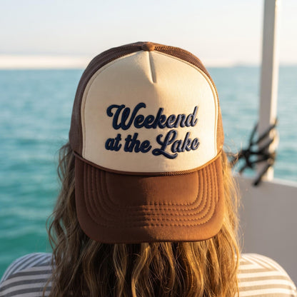 Brown and cream embroidered trucker hat reading Weekend at the Lake beside a turquoise lake