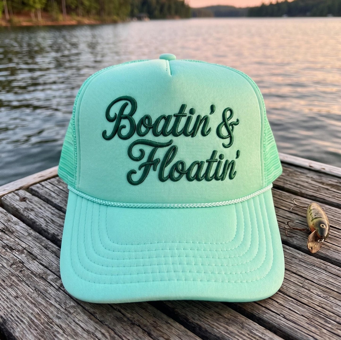 Aqua Boatin' & Floatin' embroidered trucker hat resting on a lakeside dock