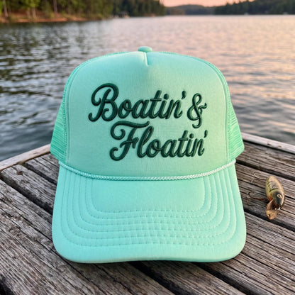 Aqua Boatin' & Floatin' embroidered trucker hat resting on a lakeside dock
