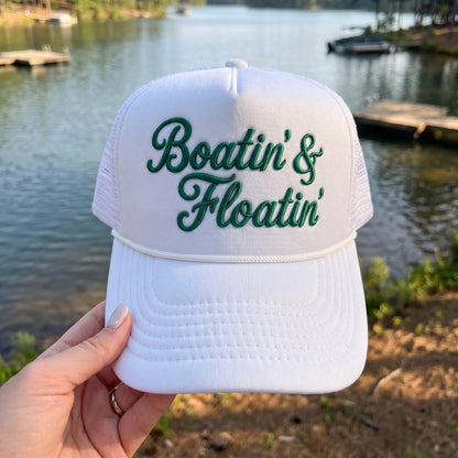 White Boatin' & Floatin' embroidered trucker hat beside a lake