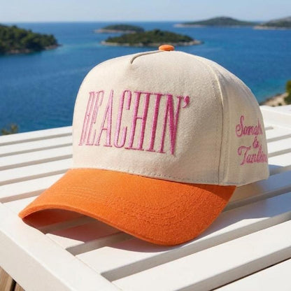 Front view of a cream and orange Beachin' embroidered trucker hat overlooking a blue coastal landscape