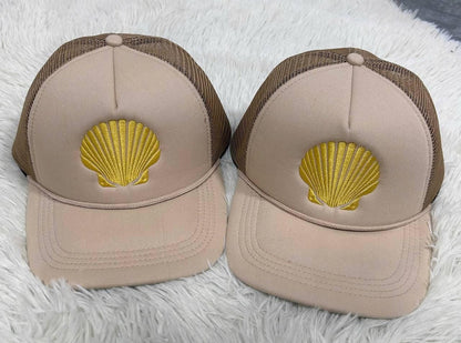Two beige trucker hats with gold seashell embroidery on a soft textured surface