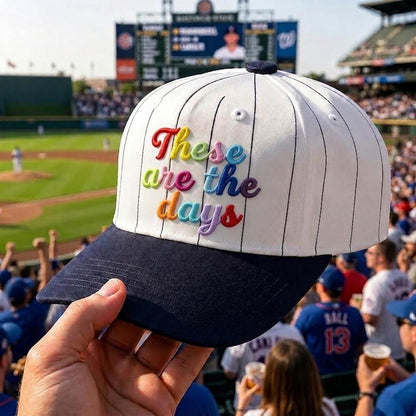 White pinstripe baseball cap with colorful “These are the days” embroidery at a baseball stadium