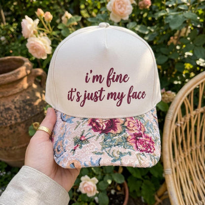 Front view of a light neutral embroidered hat with burgundy “I’m Fine, It’s Just My Face” lettering and a floral brim